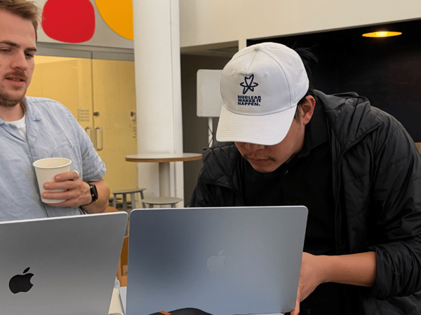 A candid indoor photograph showing two men working side-by-side on silver laptops in what appears to be a modern office or co-working space, with one man wearing a white cap featuring the text 'NUCLEAR MAKES IT HAPPEN.'