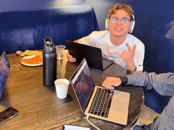 A group of people is gathered around a dark round table in a dimly lit indoor setting with deep blue velvet seating, working on laptops and sharing drinks and food.