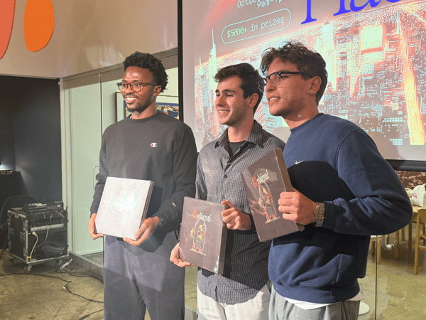 A photograph showing three young men standing side-by-side, smiling, and holding prizes or awards in front of a large projection screen displaying event details and a stylized city skyline image.