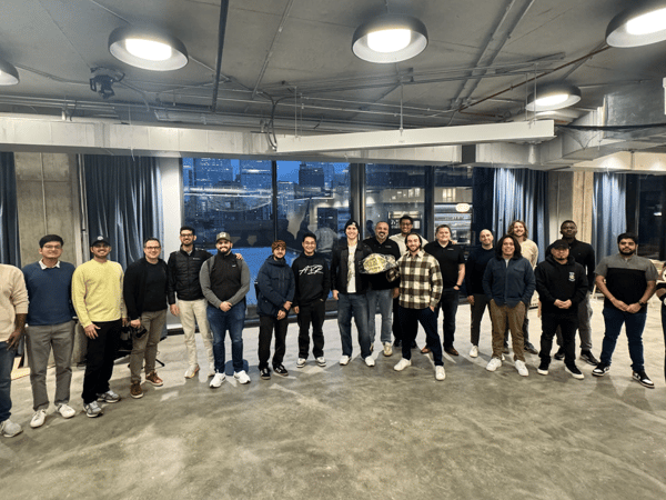 A large group of approximately 19 people, likely colleagues, pose for a group photo indoors in a modern office space with large windows overlooking a nighttime city view. One person in the center is holding a large championship belt or award.