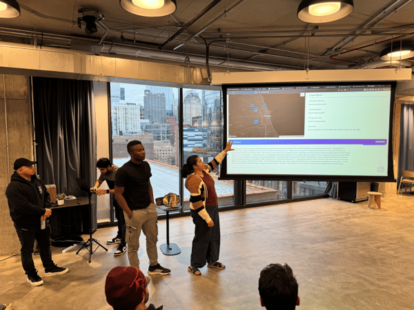 A group of people attends a presentation in a modern, high-rise venue with large windows overlooking a city skyline, likely Chicago. A woman stands pointing at a large projection screen displaying a map and data related to the Logan Square neighborhood.