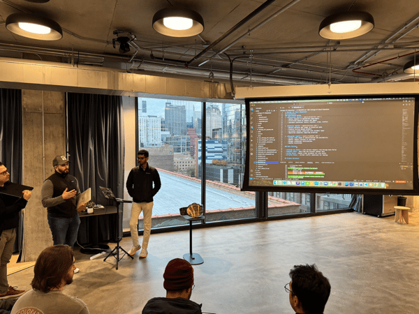 A photograph captures a technical presentation taking place in a modern office space with large windows overlooking a city skyline, featuring two presenters standing near a screen displaying code while several attendees watch from the foreground.