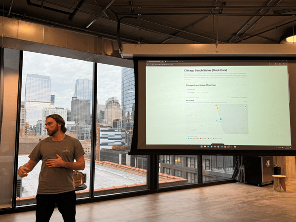 A man stands in a modern office space with large windows overlooking a city skyline, giving a presentation or demonstration displayed on a large projection screen to his right.
