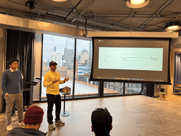 An indoor photograph captures a presentation session where a man in a yellow sweater speaks and gestures in front of a large projection screen displaying a technical diagram, with a city skyline visible through expansive windows behind him.