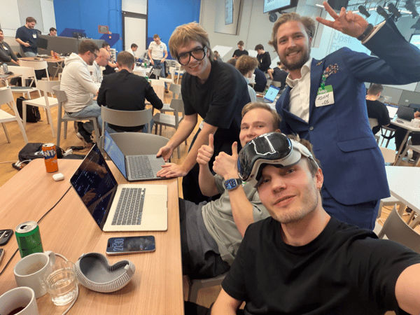 A group selfie taken by four young men at an indoor tech event or hackathon, showing them posing happily around a table with laptops and drinks while many other attendees work in the background.