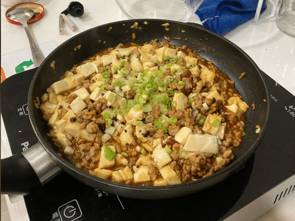 A close-up photograph of Mapo Tofu cooking in a black frying pan on a black induction cooktop, garnished generously with chopped green onions.