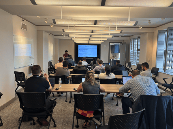 A group of people attending a technical presentation or workshop in a modern office classroom setting. A presenter stands at the front next to a large screen displaying code, while participants sit at desks with their laptops open.