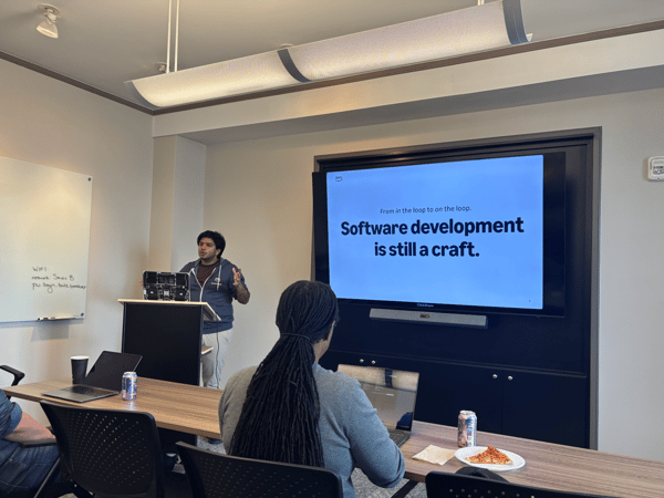 A man is giving a presentation in a conference room, with a large screen displaying text about software development while audience members sit at a table with laptops and pizza.