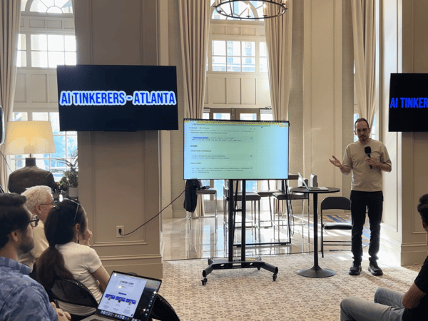 A man is giving a technical presentation titled 'AI Tinkerers - Atlanta' to an audience in a spacious, elegant indoor setting with high ceilings.