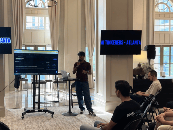 A man stands at a podium giving a presentation to an audience in a grand, high-ceilinged room with large windows. Screens in the background display 'AI TINKERERS - ATLANTA'.