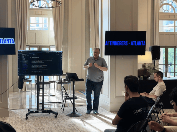A man stands in a bright, high-ceilinged room giving a presentation to a seated audience, with screens displaying 'AI Tinkerers - Atlanta'.