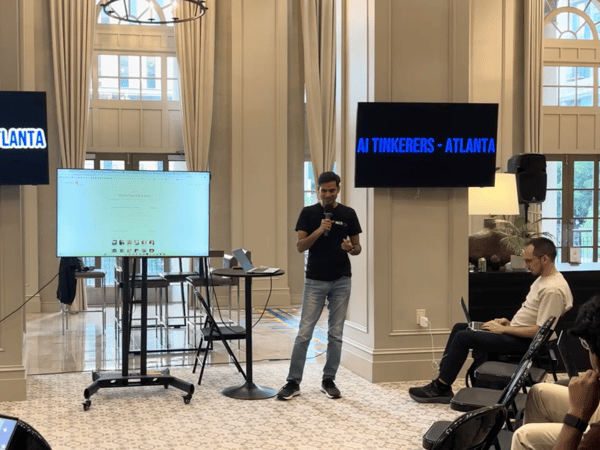 A man stands at a podium giving a presentation at an 'AI Tinkerers - Atlanta' event in a grand room with high ceilings and large windows.