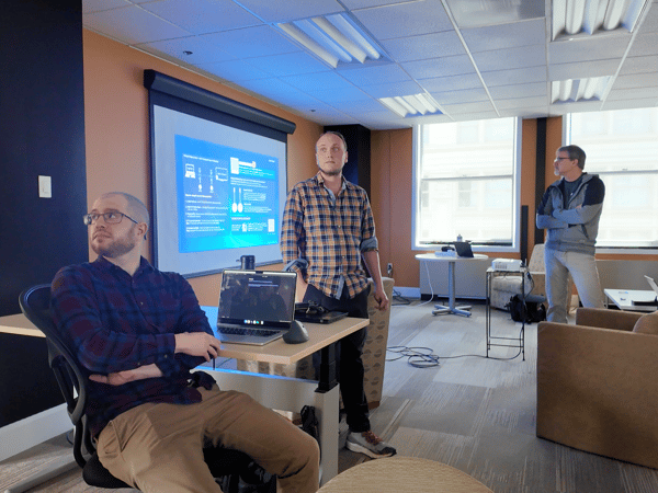 A group of men are gathered in a modern office space, focused on a technical presentation displayed on a projector screen and a laptop.