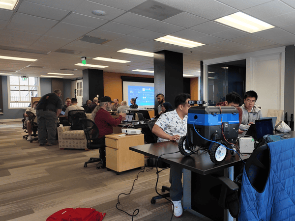 A group of people are working in a modern office or lab setting, with several individuals focused on a small, blue, wheeled robot equipped with sensors.