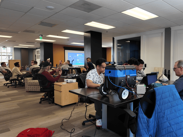 A group of people working in a modern office or lab environment, with a small blue and black robotic vehicle on a table in the foreground.