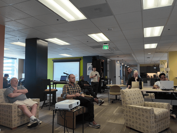 A group of people are gathered in an open-plan office space, some sitting in armchairs and others at desks, facing a projector screen in the background.