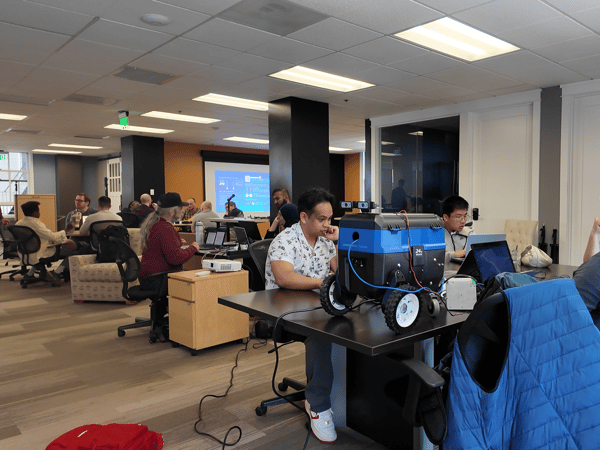 A group of people are working in a modern office or lab environment, centered around a table with a blue robotic device and several open laptops.