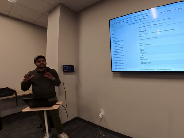 A man in a green hoodie and cap is giving a presentation next to a laptop stand, gesturing with his hands while a large wall-mounted screen displays a GitHub README page for 'Cognitive-SRE'.