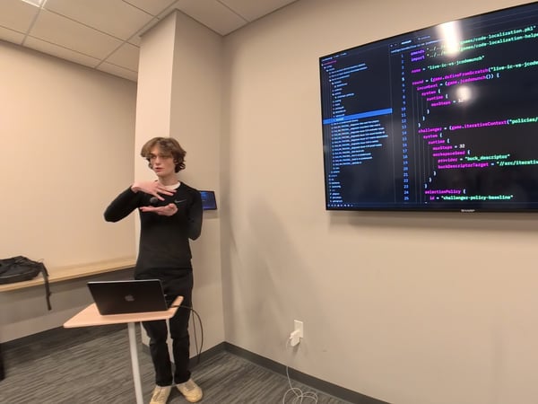 A young man stands in an office or classroom setting, gesturing with his hands while presenting code displayed on a large wall-mounted monitor.