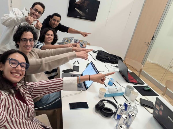 A group of five smiling young people, likely colleagues or students, pose for a selfie around a cluttered white table, all pointing their arms forward in unison.