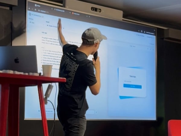 A man is giving a technical presentation indoors, standing in front of a large screen displaying development instructions and a web application interface labeled 'Todo App,' while holding a microphone and pointing at the content.