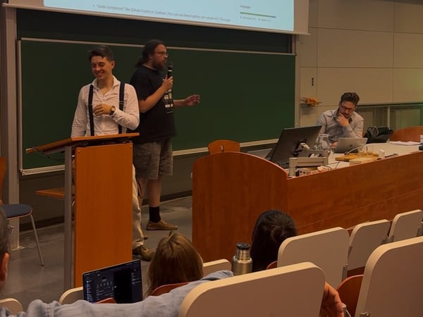 A low-light photograph taken inside a lecture hall shows two men standing near a lectern, one speaking into a microphone, while a third man is seated at a table to the right, working on a laptop. A presentation slide is visible on the screen behind them, featuring text about code completion tools.