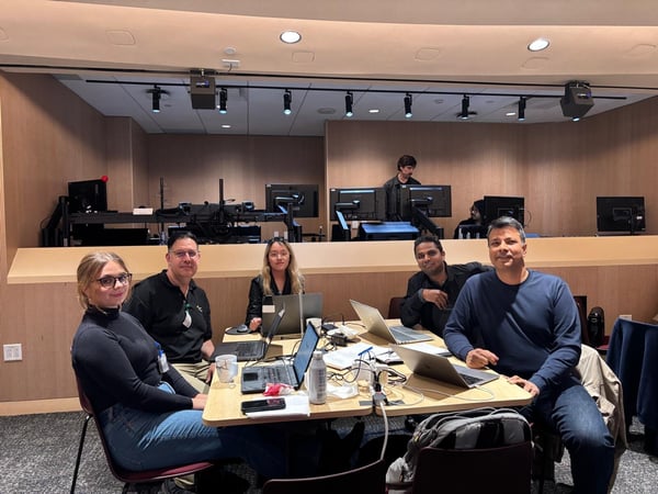 A group of six colleagues is gathered around tables in a modern indoor setting, likely an office or control room, working on laptops and smiling at the camera. Several computer monitors and equipment racks are visible on a raised platform behind them.