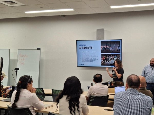 A photograph captures an indoor presentation or workshop where a woman stands near a large screen displaying a slide titled 'WHO WE ARE AI TINKERERS' while speaking to an audience seated at tables.