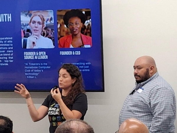 A photograph captures an indoor presentation where a woman is speaking and gesturing in front of a large screen displaying a slide about 'AI Tinkerers' and its founders. A man stands beside her, observing, while audience members sit in the foreground.