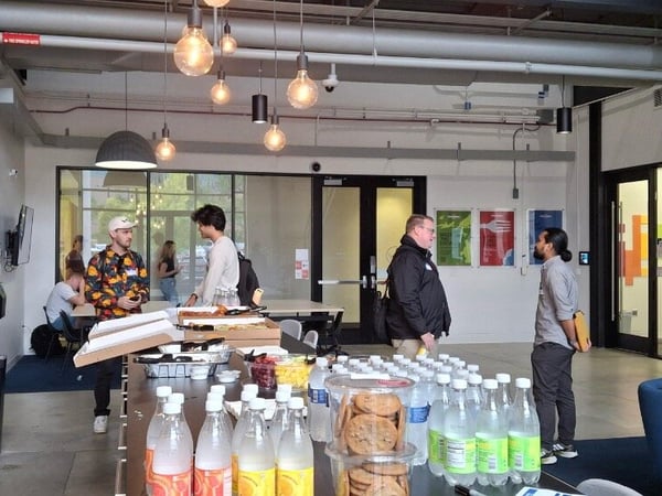 A photograph captures an indoor social gathering in a modern office or communal workspace, where attendees are standing around a long counter laden with bottled drinks and snacks.