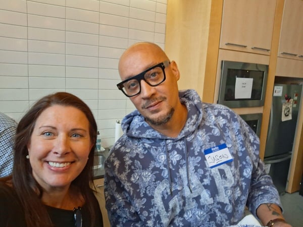 A close-up photograph of two people, a smiling woman and a man wearing large black glasses and a floral hoodie, posing together indoors in what appears to be a modern office kitchen or breakroom.