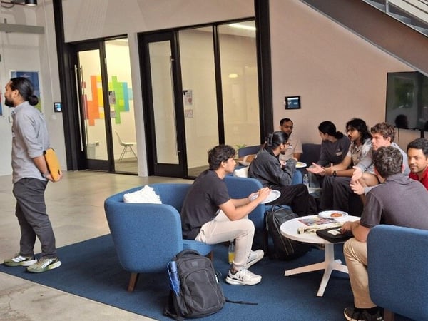 A group of approximately eight people is gathered in a modern, indoor common area or office lobby, sitting in blue lounge chairs around a white coffee table, engaged in discussion and eating snacks. A staircase with metal railings is visible on the right, and posters are displayed on the wall to the left.
