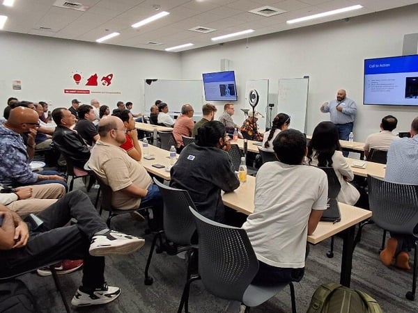 A photograph capturing a large group of people attending a presentation or workshop inside a modern conference room, with presenters standing near screens displaying slides.
