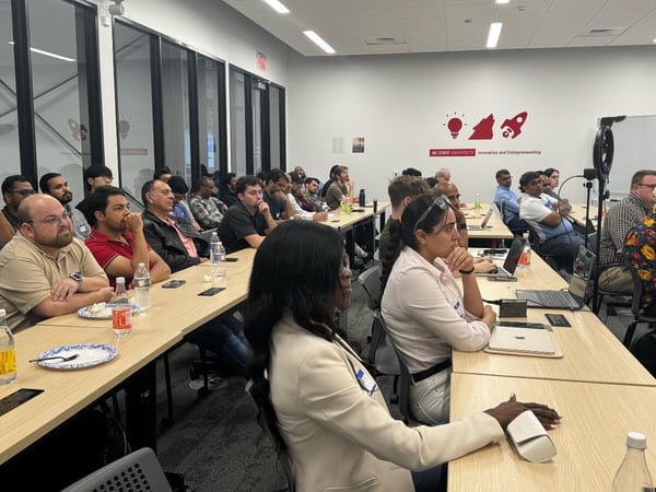A photograph capturing a large group of people seated at long tables in a modern, well-lit conference room, attending a presentation or lecture. The back wall features a red decal for NC State University Innovation and Entrepreneurship.