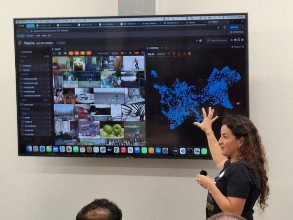 A woman is giving a technical presentation indoors, pointing at a large monitor displaying a dark-mode interface with a grid of images and a large blue point cloud data visualization.