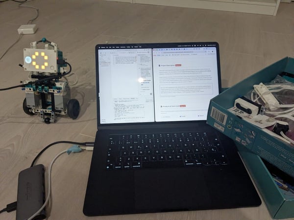 A photograph showing a robotics project setup on a light wooden floor, featuring a small, assembled robot next to an open laptop displaying code and project documentation.