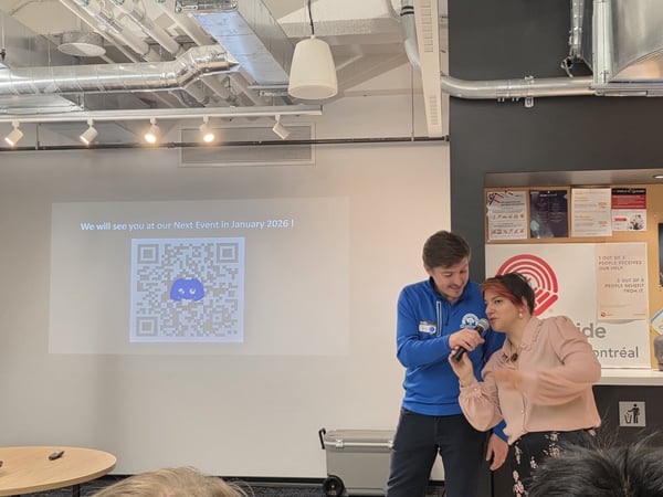 A candid photograph taken indoors showing a man and a woman speaking into a microphone during an event, standing in front of a projected screen displaying a Discord QR code and event information.