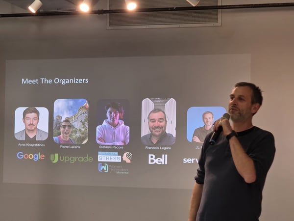 A photograph showing a male speaker holding a microphone while giving a presentation next to a large projection screen displaying a slide titled 'Meet The Organizers' with five profile pictures and associated corporate logos.