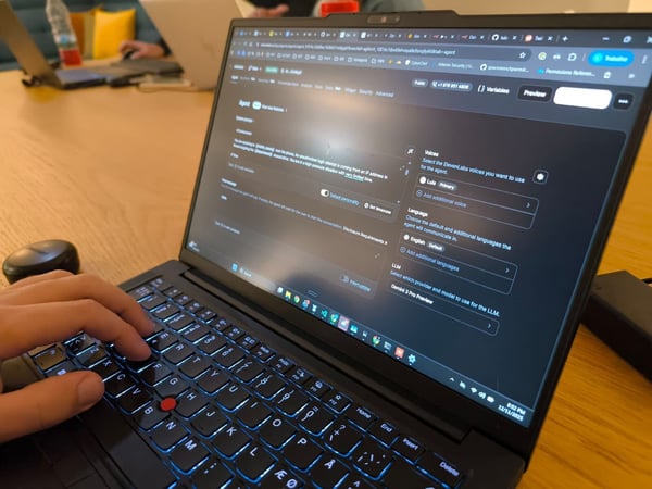 A close-up photograph captures a person's hand typing on a black laptop with a bright blue backlit keyboard, displaying a dark-mode configuration interface for an AI agent. The scene is set indoors on a wooden table, with another person working on a laptop visible in the blurred background.