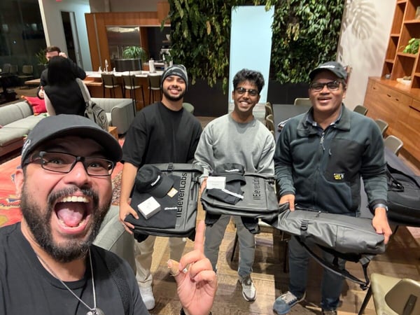 A group selfie captures four men indoors, smiling and posing while holding dark backpacks and merchandise branded with '11ElevenLabs,' set against a backdrop of a modern office lounge featuring a vertical living green wall.