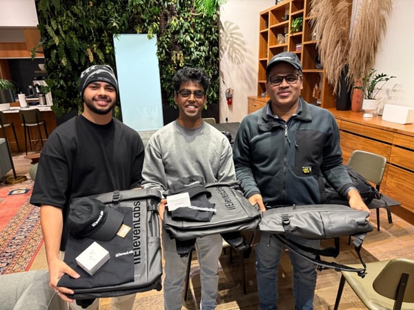 Three men stand indoors, smiling and posing while holding black bags and merchandise branded with "ElevenLabs" in front of a vertical garden wall and wooden shelving.