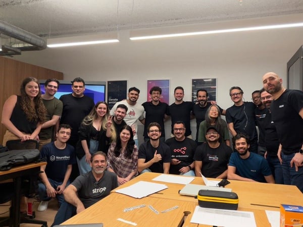 A large group of approximately twenty people, likely colleagues, pose together for a team photograph indoors in a modern office space, gathered around a wooden conference table.