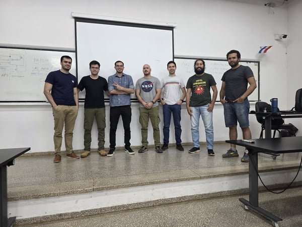 A group of seven men posing for a photo in front of a projector screen and a whiteboard in a classroom or office setting.