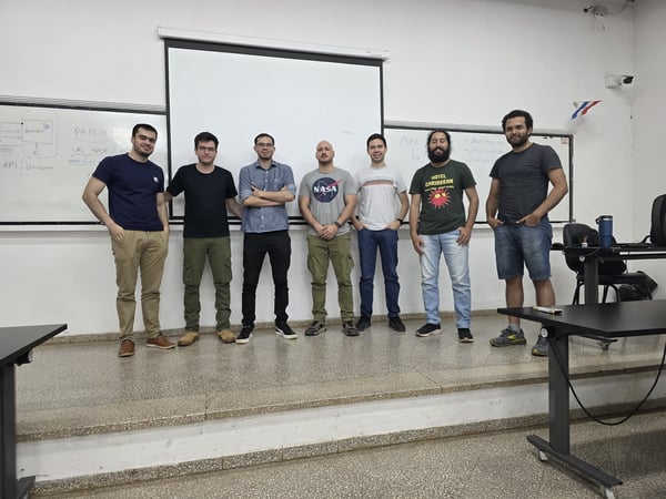 A group of seven men standing in a row for a photo in front of a whiteboard and a blank projection screen in a classroom or lab setting.