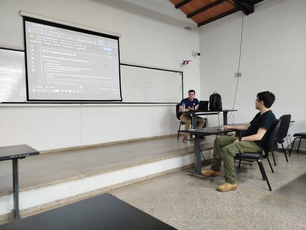 A man is giving a presentation in a classroom, with a projector screen showing a Google Colab notebook in Spanish about 3D object generation.