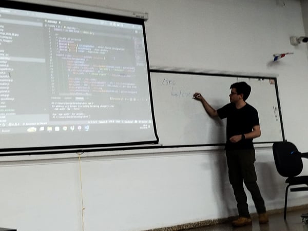 A man stands in a classroom or lecture hall, writing on a whiteboard while a projector displays JavaScript code on a screen next to him.