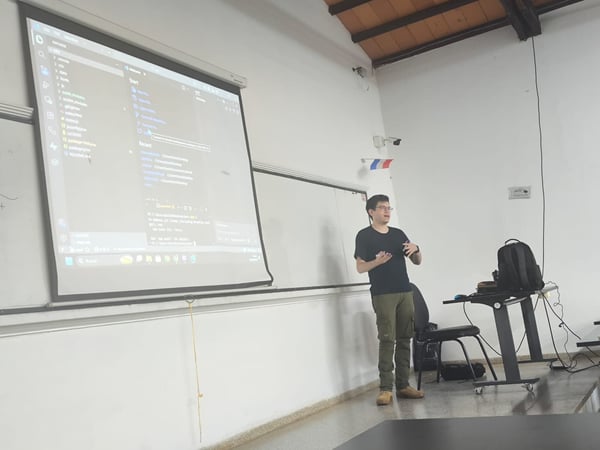 A man is giving a presentation in a classroom, standing next to a large projection screen that displays a code editor.