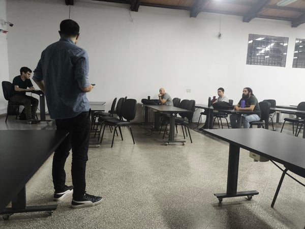 A man stands in the foreground of a classroom-like setting, facing a small group of people seated at tables.