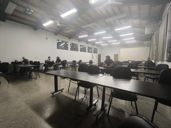 A wide-angle photograph of a sparsely populated classroom featuring rows of dark tables and chairs under a slanted ceiling with bright fluorescent lights.