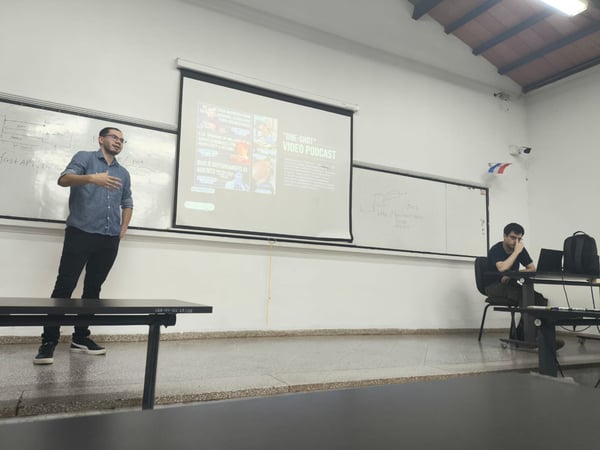 A man stands in front of a projector screen giving a presentation in a classroom setting, while another person sits at a desk with a laptop to the right.